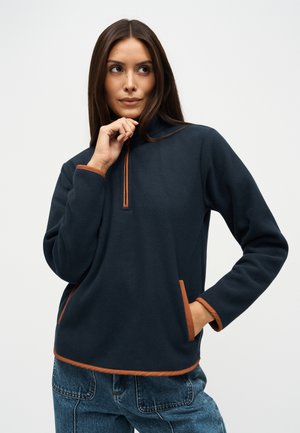 Une femme pose sur un fond blanc uni, vêtue d’un pull en polaire bleu marine avec des finitions couleur rouille et d’un jean bleu.