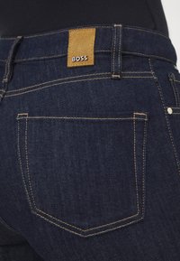 Jeans de mezclilla azul oscuro con costuras amarillas, que presentan un parche de ante marrón etiquetado como "BOSS" en la cinturilla y dos bolsillos traseros.