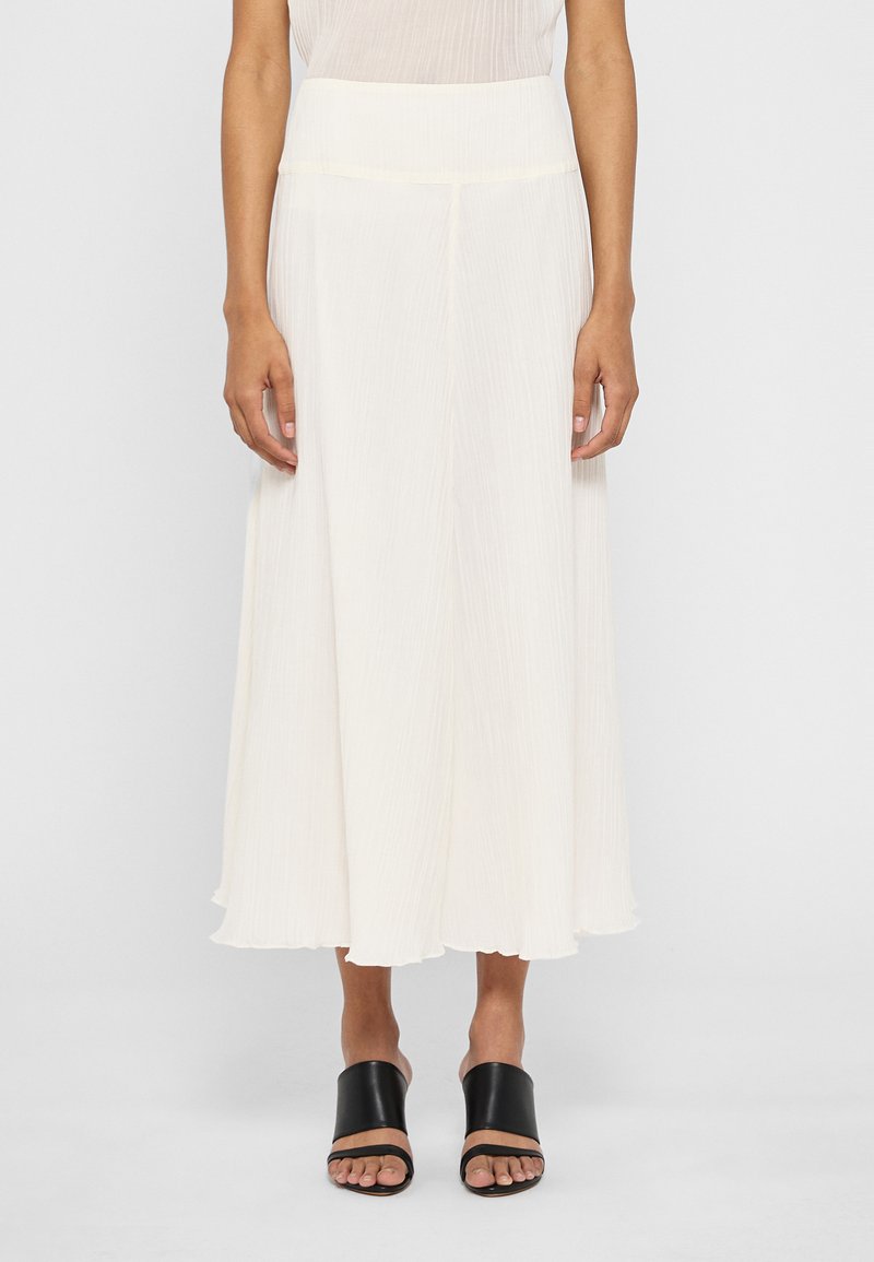 Jupe maxi plissée blanche avec une taille haute, une silhouette fluide et une texture douce, associée à des sandales noires élégantes.