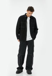Camisa de mezclilla negra con dos bolsillos en el pecho, combinada con pantalones cargo negros que cuentan con múltiples bolsillos. Camiseta blanca debajo. Tejido texturizado.