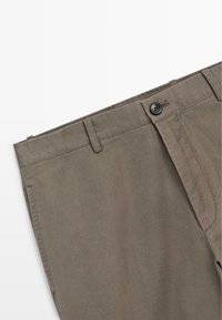 Zbliżenie na brązowe spodnie pokazujące pas z pętlami na pasek, zapięcie na guzik oraz część przedniej kieszeni na białym tle.