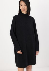 Čierne pletené šaty so vysokým výstrihom, širokými rukávmi a bočnými vreckami. Majú jemnú textúru a uvoľnený, oversized strih.