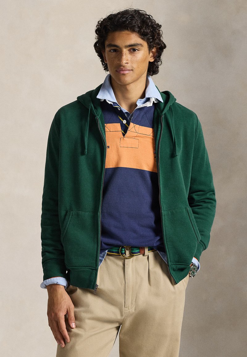 Sweatshirt verde com fecho de correr e dois bolsos frontais, usado sobre um polo às riscas laranja e azul-marinho, e calças bege. Detalhes notáveis incluem uma gola azul e um cinto às riscas.