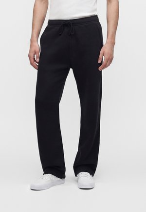 Man draagt zwarte broek met trekkoord en witte sneakers en staat tegen een effen witte achtergrond.