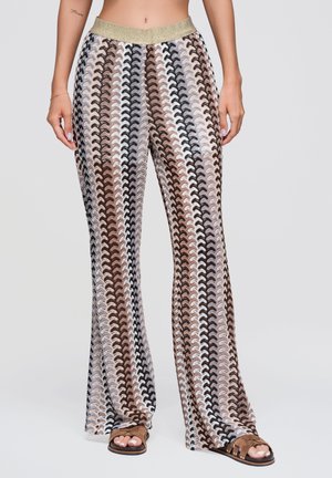 Femme portant un pantalon taille haute à jambes larges avec un motif ondulé marron, noir et beige et une ceinture élastique dorée, associé à des sandales ouvertes marron.