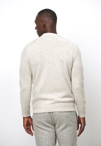 Helle beige Strickjacke mit einem gesprenkelten Muster, Rundhalsausschnitt, gerippten Bündchen und Saum. Getragen mit grauen strukturierten Hosen.