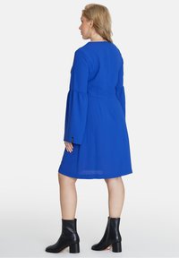Königsblaues Kleid mit langen, glockenförmigen Ärmeln, einem Reißverschluss hinten und einem ausgestellten Saum. Kombiniert mit schwarzen Knöchelstiefeln. Glatte Stofftextur.