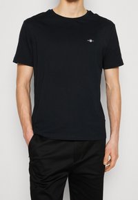 T-shirt di cotone nero con scollo a girocollo e maniche corte, con un piccolo logo bianco sul petto a sinistra. Texture liscia, vestibilità regolare.