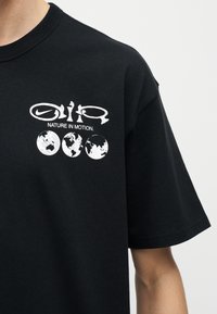 Zwart T-shirt met witte grafische opdruk, inclusief Nike-logo, gestileerde tekst "NATURE IN MOTION" en drie wereldbolillustraties op de borst.