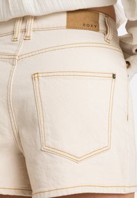 Weiße Denim-Shorts mit kontrastierenden gelben Nähten, ausgestattet mit einer kleinen Gesäßtasche und einem hellbraunen Roxy-Label am Bund.