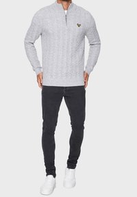 Pull en maille torsadée gris avec un col demi-fermeture éclair et un petit logo jaune. Associé à un jean noir slim et des baskets blanches.