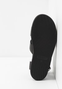 Sandale noire avec une semelle en caoutchouc texturé, des sangles larges et un semelle profilée. Présente un motif ondulé et une empreinte de logo sur la semelle.