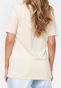 Krémové tričko s krátkým rukávem a volnějším střihem, vyrobené z měkkého materiálu. Má kulatý výstřih a hladkou texturu, s mírně delší zadní částí.