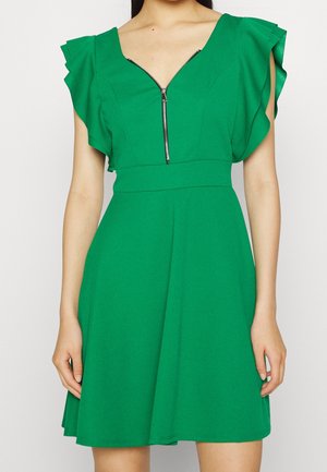 Grünes Kleid mit kurzen Rüschenärmeln, V-Ausschnitt mit Reißverschluss vorne, tailliertem Schnitt und ausgestelltem Rock, getragen von einer Person.