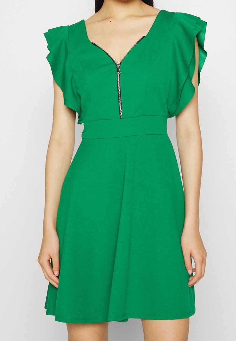 Robe verte avec manches courtes à volants, encolure en V avec fermeture éclair frontale, taille ajustée et jupe évasée portée par une personne.