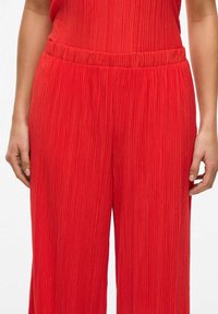Pantalones rojos con pliegues, de pierna ancha, que cuentan con una cinturilla elástica y un tejido de textura vertical en toda la tela.