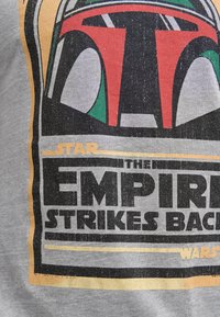 Grå t-shirt med et vintage-inspireret tryk af Boba Fetts hjelm, med røde og grønne accenter, fed sort tekst og en slidt finish.