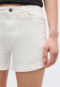 Κοντινό πλάνο ατόμου που φορά λευκά τζιν σορτς με κουμπί μπροστά και εμφανείς πλαϊνές τσέπες, το χέρι ακουμπά κοντά στο τελείωμα πάνω στον μηρό.