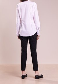 En vit och ljusrosa randig långärmad skjorta med rundad nederkant, tillsammans med smala svarta byxor och svarta loafers, sedd bakifrån.