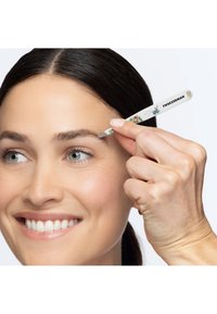 Femme souriante utilisant une pince à épiler à motif floral de Tweezerman pour façonner son sourcil sur un fond blanc.