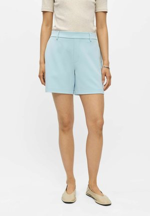 Femme portant un short bleu clair sur mesure, un top beige à manches courtes et des chaussures plates beiges, debout devant un fond uni.