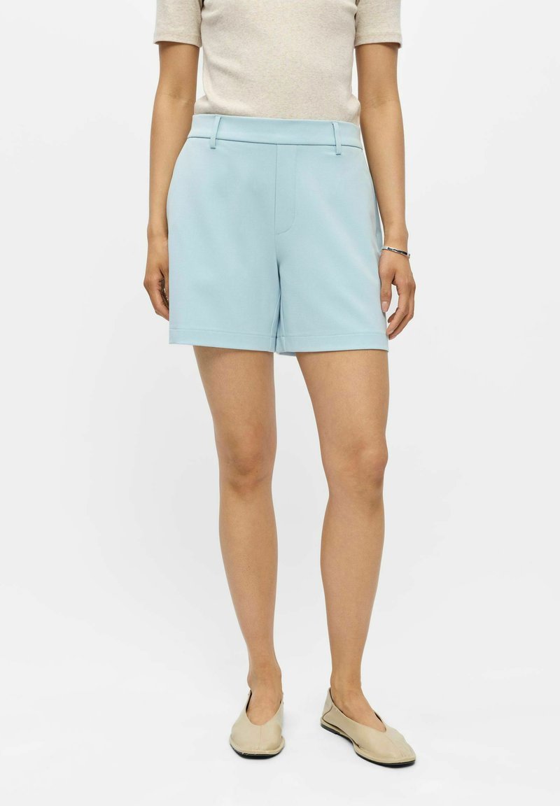 Femme portant un short bleu clair sur mesure, un top beige à manches courtes et des chaussures plates beiges, debout devant un fond uni.