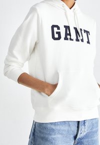 Persona con cappuccio bianco con il testo "GANT" sul petto e jeans in denim leggero blu, mani infilate nella tasca del cappuccio.