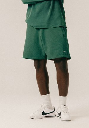 Person iført mørkegrønne shorts og hvide Nike-sneakers med hvide crew-sokker, stående foran en neutral baggrund.