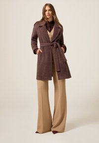 Donker mauve jas met een textuurafwerking, ceintuur in de taille, grote revers en zijzakken, gecombineerd met lichtbeige wijde broekspijpen.