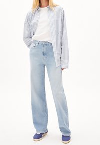 Kvinde iført lyseblå vide ben jeans, hvid t-shirt, blåt stribet skjorte og lilla sneakers står op ad en hvid baggrund.
