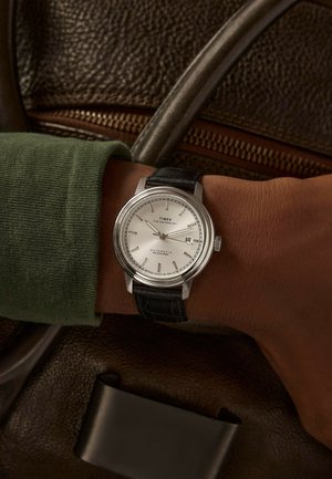 Montre analogique argentée avec un cadran blanc, un bracelet en cuir noir et une fonction date. Présentée sur un poignet portant un pull vert, près d'un sac en cuir marron.