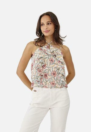 Mujer con blusa halter sin mangas con volantes y estampado floral, con tirantes cruzados y pantalones blancos de cintura alta, de pie con las manos detrás de la espalda.