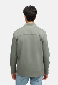 Grüne strukturierte Jacke mit Kragen und langen Ärmeln, mit einem Rückenteil im Schulterbereich, kombiniert mit hellblauen Jeans.