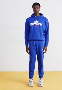 Kék Nike kapucnis pulóver "sportswear" felirattal, illeszkedő kék melegítőnadrággal és fehér sportcipőkkel. Sima anyag, laza fazon és bordázott mandzsett�ák.