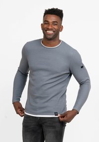 Graues langärmliges Ripp-Shirt mit rundem Halsauschnitt und weißem Besatz, versehen mit einem kleinen schwarzen Etikett am Saum. Getragen mit dunklen Jeans.