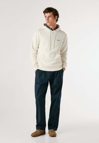 Lichte crèmekleurige hoodie met een voorzak en een trekkoord, gedragen met donkerblauwe broek; beige schoenen met witte strepen maken de outfit af.
