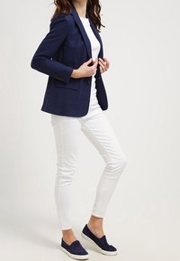 Blazer bleu marine, t-shirt blanc, jean skinny blanc et chaussures slip-on bleues marines. Le blazer présente des revers crantés et deux boutons.