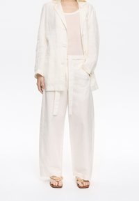 Ľanový blazer a široké nohavice, oboje v odtieňoch off-white, v kombinácii s béžovými sandálmi so textúrovaným kvetinovým akcentom. Jednoduchý, uvoľnený strih.