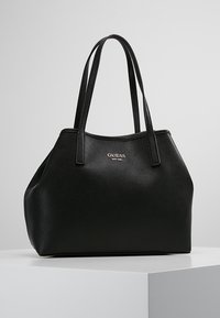 Sac en toile noir en cuir texturé de la marque Guess avec deux anses et un logo doré, posé sur un piédestal blanc contre un fond gris.