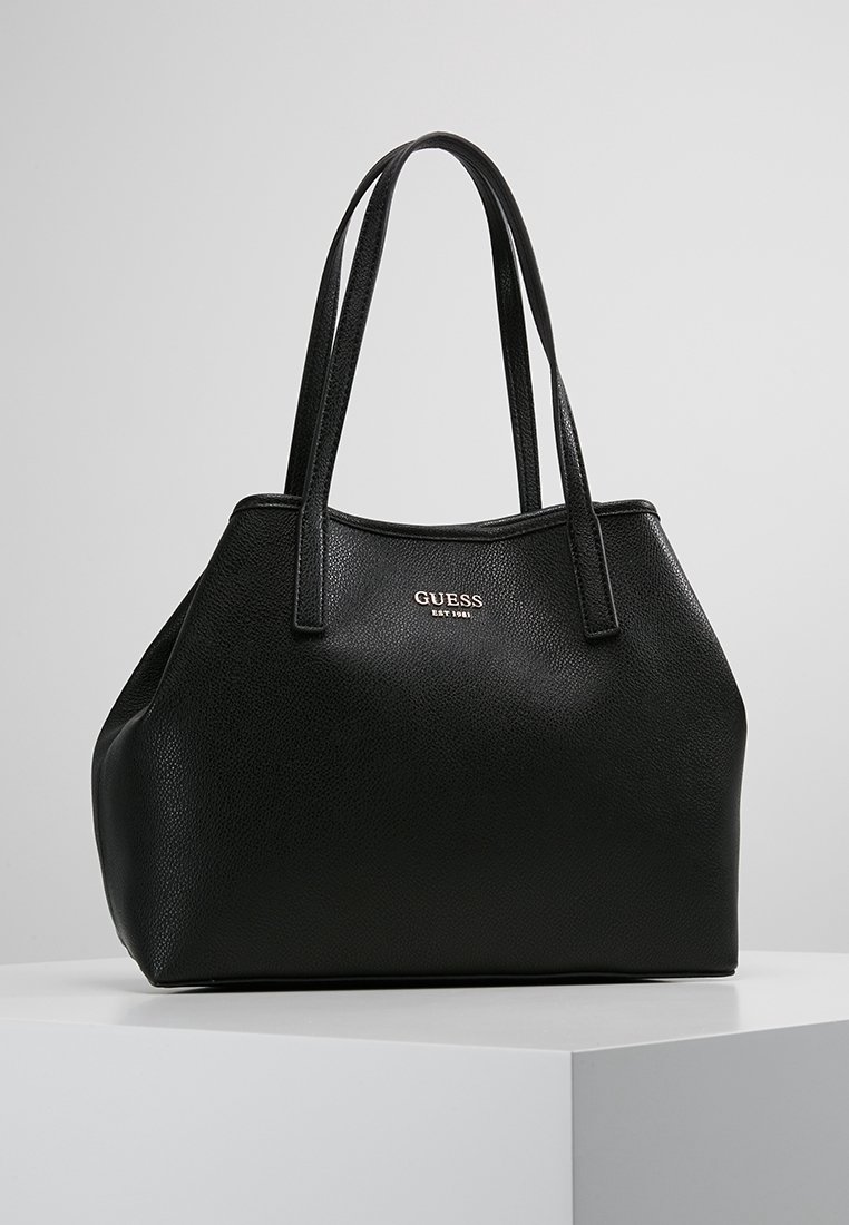 Sac en toile noir en cuir texturé de la marque Guess avec deux anses et un logo doré, posé sur un piédestal blanc contre un fond gris.