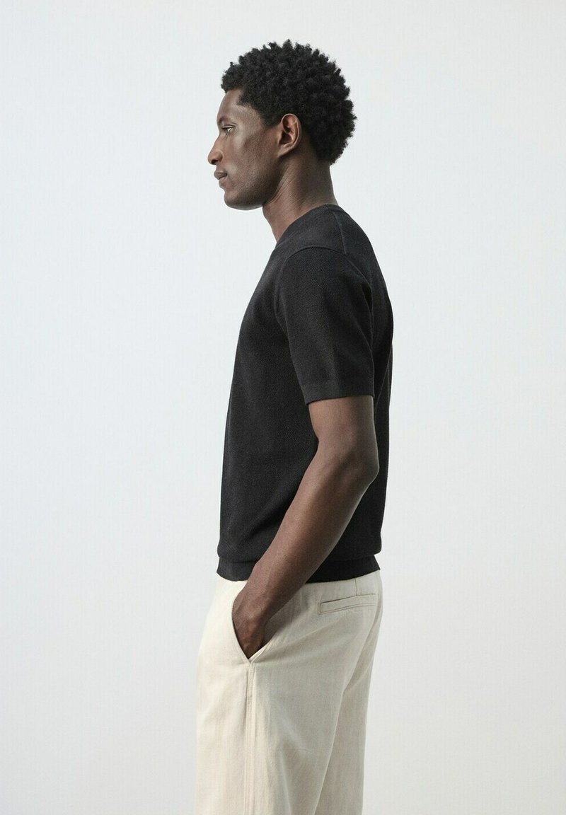 Homme debout de profil, les mains dans les poches, portant une chemise noire à manches courtes et un pantalon de couleur claire, sur un fond uni.