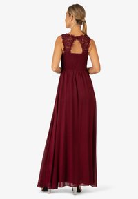 Burgunderrotes Maxikleid mit floralem Spitzenrücken, geraffter Taille und fließendem Rock. Es hat kurze Ärmel und eine durchsichtige Textur. Klare Absätze sind sichtbar.