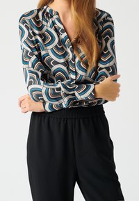 Blouse à motifs en noir, beige, bleu et orange. Dotée d'un col rond et de manches longues. Associée à un pantalon noir à taille élastique.
