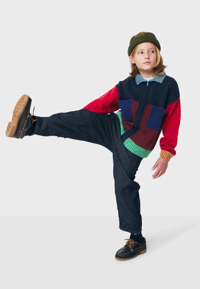 Mehrfarbiger gestrickter Pullover mit roten, blauen und grünen Abschnitten; schwarze gestreifte Hose; schwarze Schuhe mit braunen Sohlen; grüner Beanie.