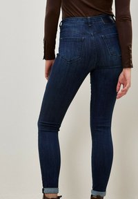 Jeans ajustados de color azul oscuro con un lavado claro, cintura de altura media y dobladillos con vuelta; presentan dos bolsillos traseros y una etiqueta de cuero con marca.