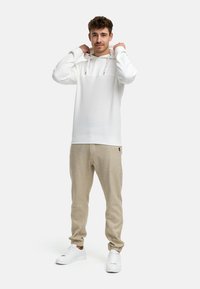 Witte hoodie met trekkoorden, textuur gebreid ontwerp, gecombineerd met lichtbeige tapered broek en witte sneakers, staand op een effen achtergrond.