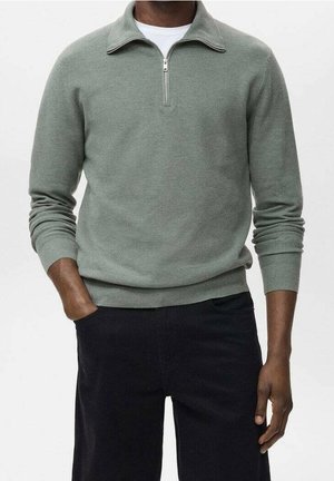Homme portant un pull gris clair à fermeture éclair quart de zip sur une chemise blanche avec un pantalon noir, une main dans la poche, devant un fond clair uni.