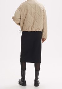 Osoba w beżowej pikowanej kurtce, czarnej prążkowanej spódnicy midi, przezroczystych czarnych rajstopach i czarnych masywnych botkach za kostkę, stojąca na białym tle.