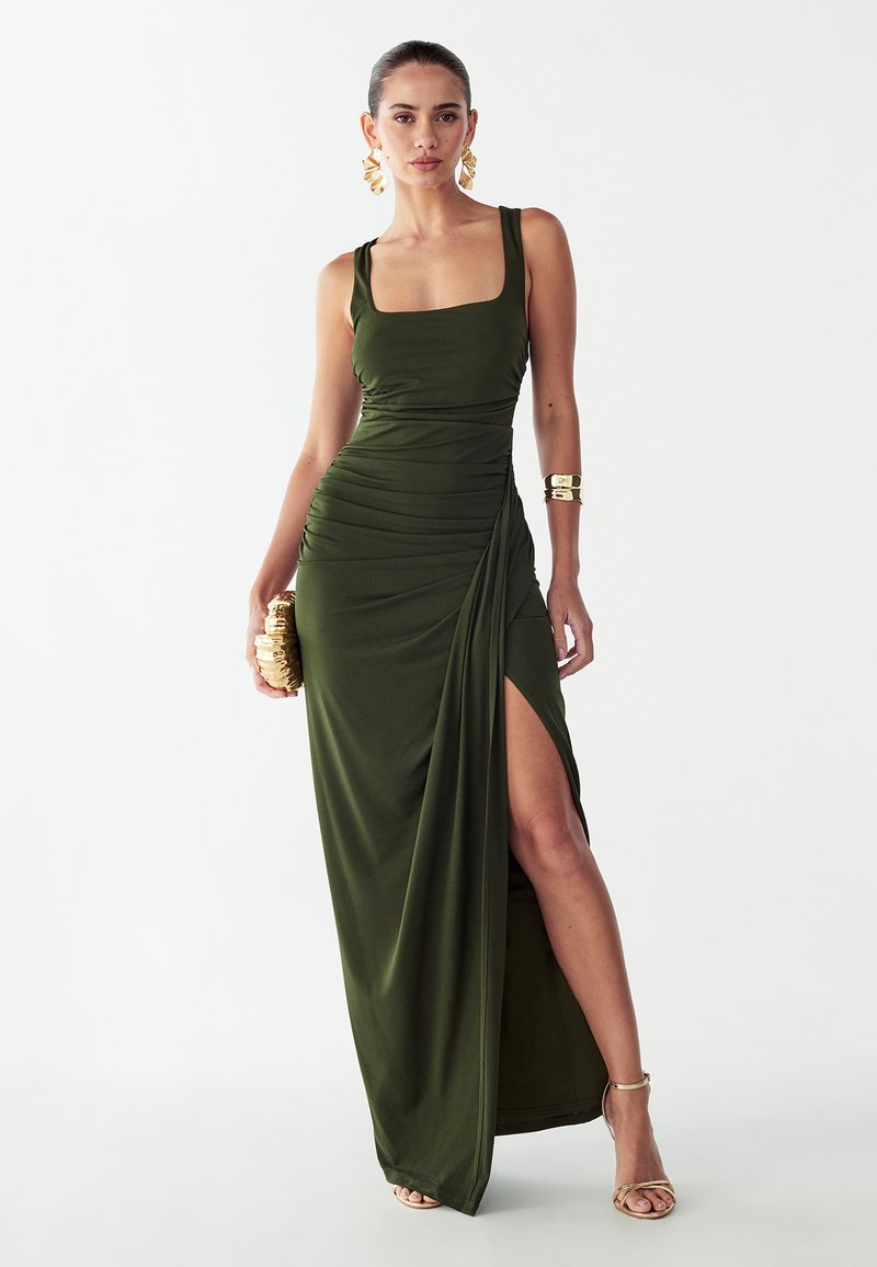 Olijfgroene, vloerbedekkende jurk met een aangesloten lijfje, een vierkante halslijn, plooien aan de zijkant en een hoge split. Gecombineerd met gouden accessoires.