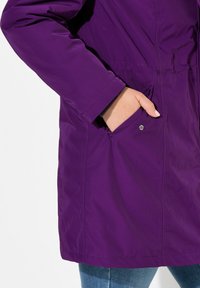 Giacca impermeabile viola con design aderente, vita elastica e tasche laterali. Il materiale ha una texture liscia con un leggero lucido.